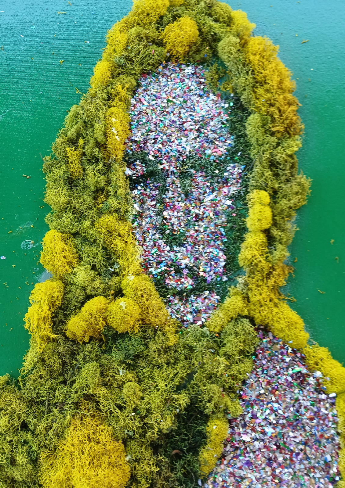 A reflective mosaic face surrounded by green moss against a turquoise ground